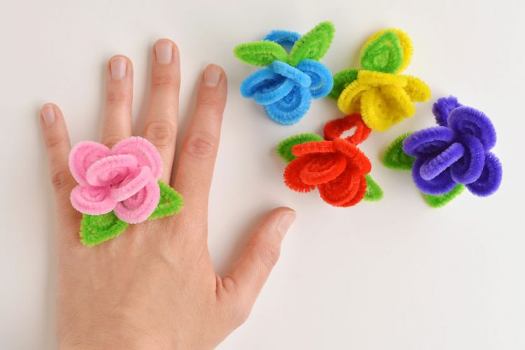A hand wearing a pink flower ring, with four other colours on the table beside the hand