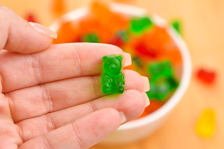 A single green gummy bear held above a bowl of gummy candy