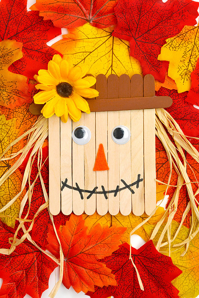 Scarecrow popsicle stick craft on a background of fall leaves